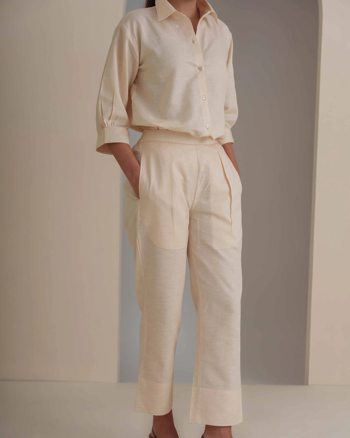 A model wearing a matching light cream linen two-piece set consisting of a button-down shirt with rolled sleeves and high-waisted cropped trousers with large patch pockets.
