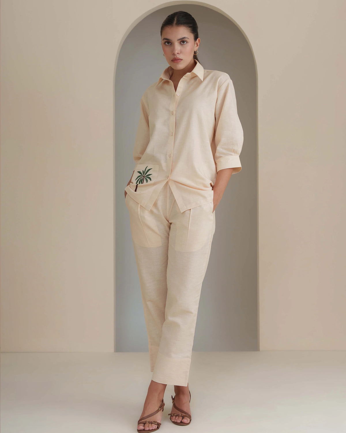 A woman models a matching cream-colored linen shirt and trousers set with a small green palm tree embroidery on the shirt pocket, set against a minimalist beige background with an arched doorway.