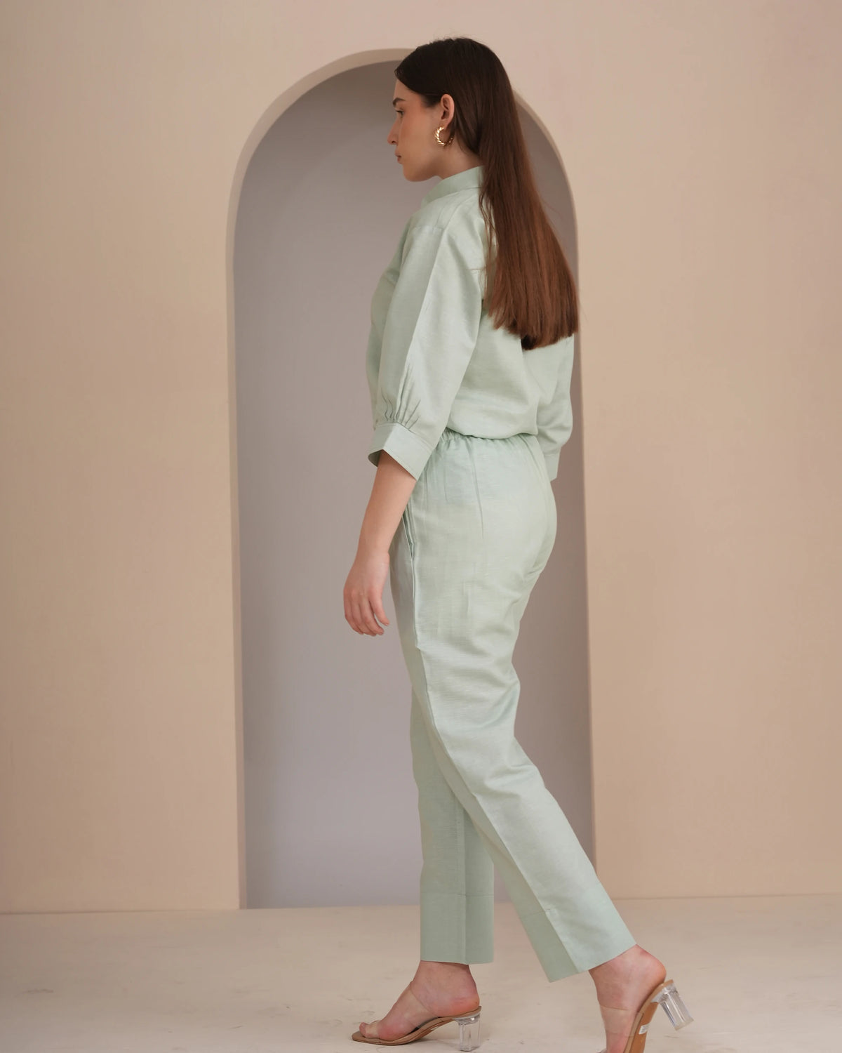 A woman wearing a light sage green two-piece set consisting of a long-sleeve collared top and matching cropped trousers, standing in profile against a beige studio backdrop with an arched alcove.