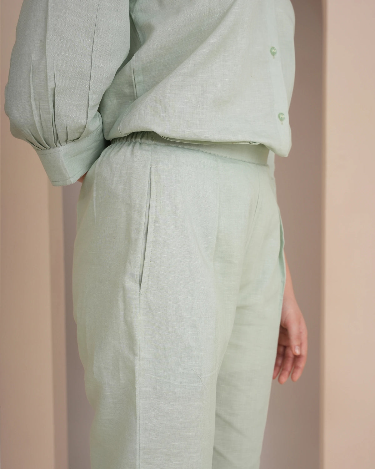 A close-up of a person wearing a light sage green linen two-piece set, featuring a long-sleeve button-up shirt and matching pleated trousers, set against a soft beige background.