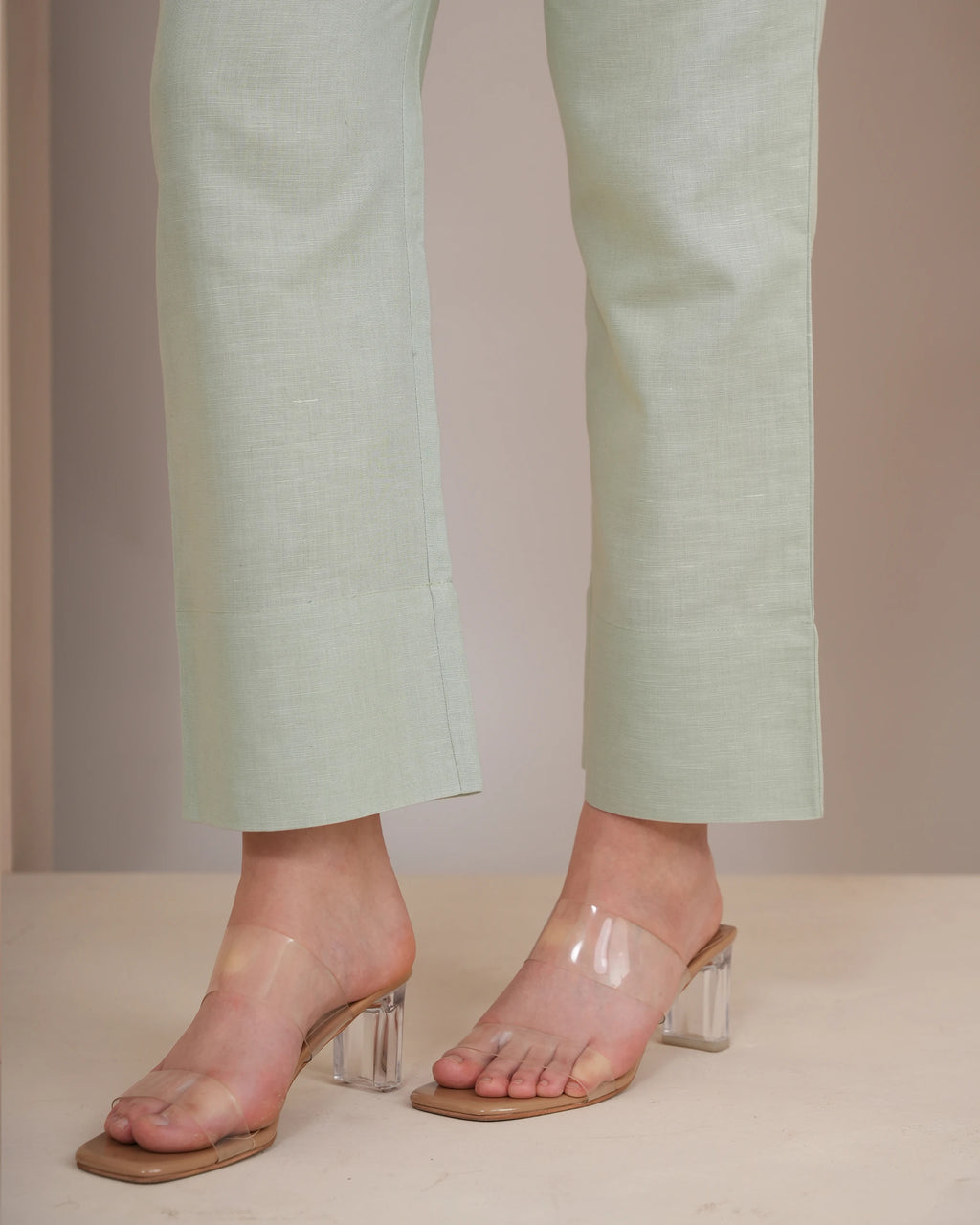 A close-up of a person's feet wearing pale sage green cropped trousers and clear strap mule sandals with chunky clear block heels, standing against a neutral beige background.