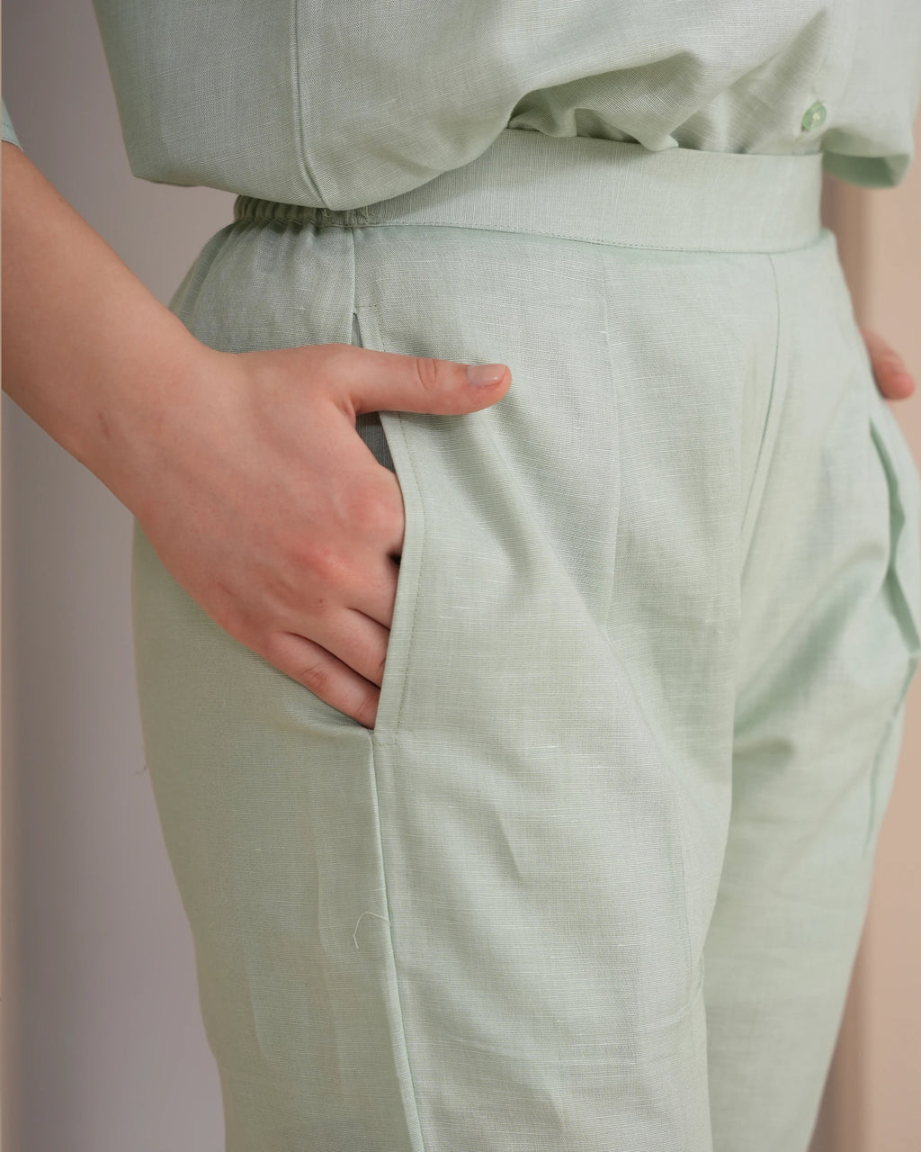 Close-up of a person wearing light sage green linen trousers with a side pocket, shown with one hand in the pocket.