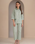 A woman stands in a studio wearing a light sage green matching set consisting of a long-sleeve button-up shirt with a small black palm tree embroidery on the pocket and matching wide-leg cropped pants.