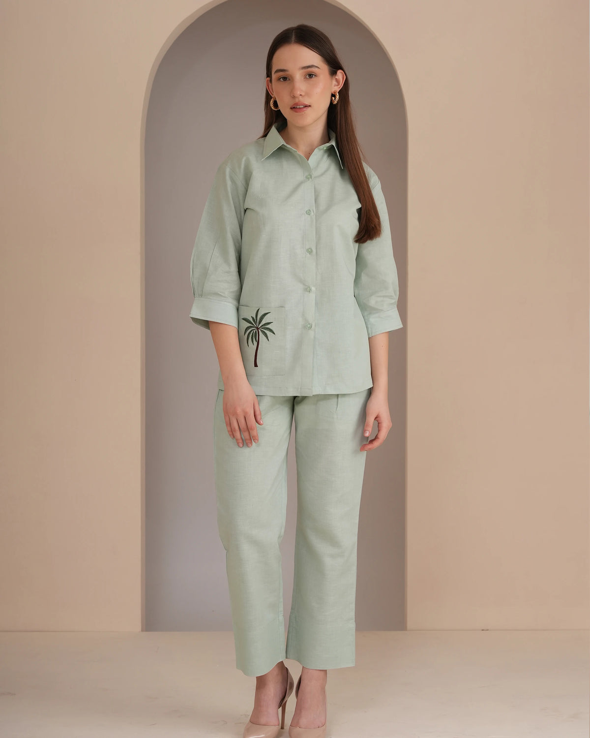 A woman stands in a studio wearing a light sage green matching set consisting of a long-sleeve button-up shirt with a small black palm tree embroidery on the pocket and matching wide-leg cropped pants.