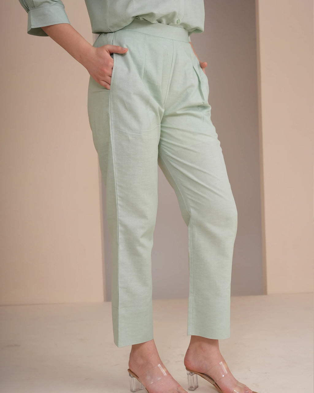 A person wearing light sage green cropped trousers with a matching top and clear high-heeled sandals, standing against a soft beige studio backdrop.