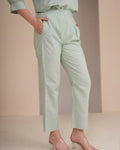 A person wearing light sage green cropped trousers with a matching top and clear high-heeled sandals, standing against a soft beige studio backdrop.