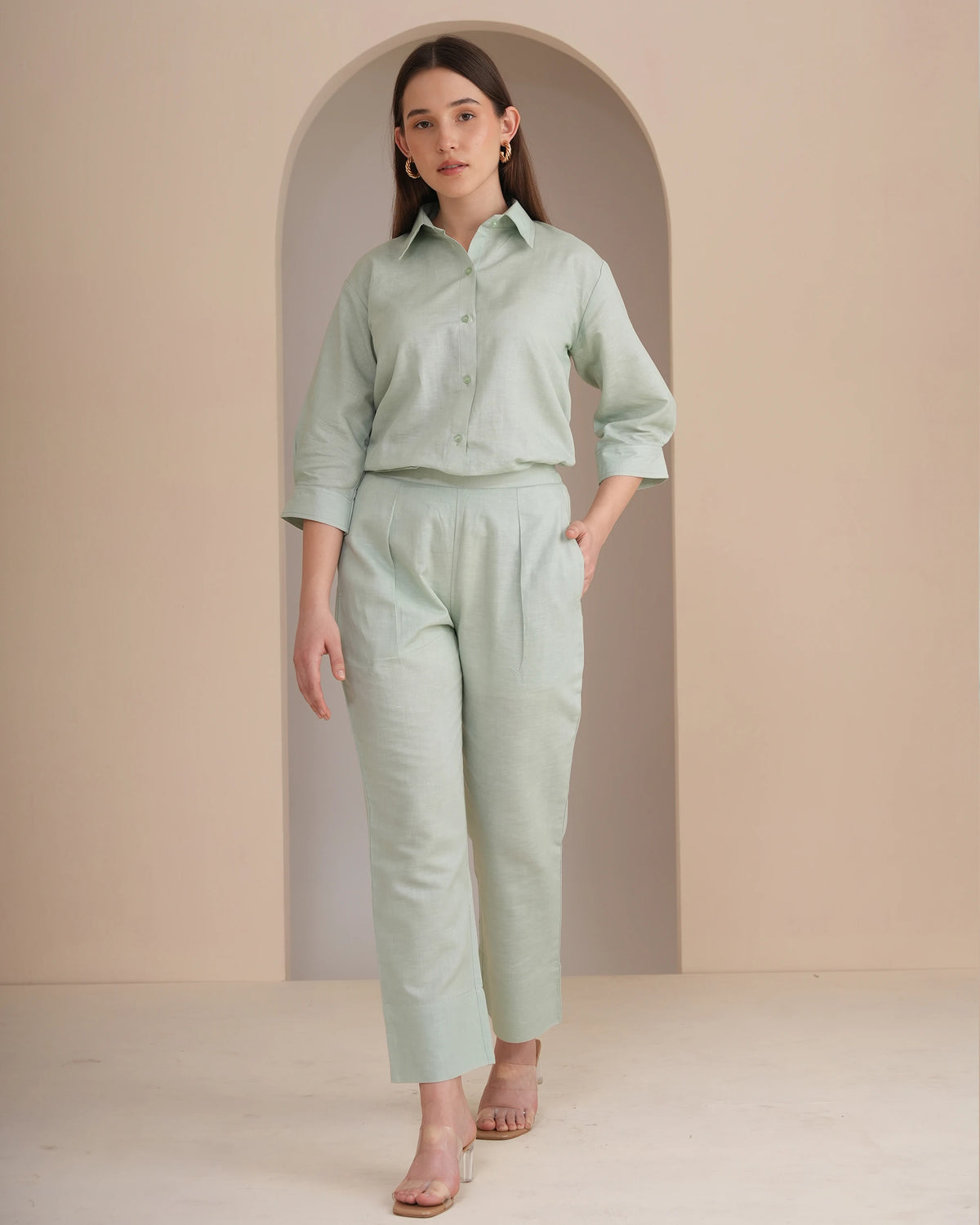 A woman models a light sage green two-piece linen set consisting of a button-down shirt and matching cropped trousers, standing in front of a beige arched wall.