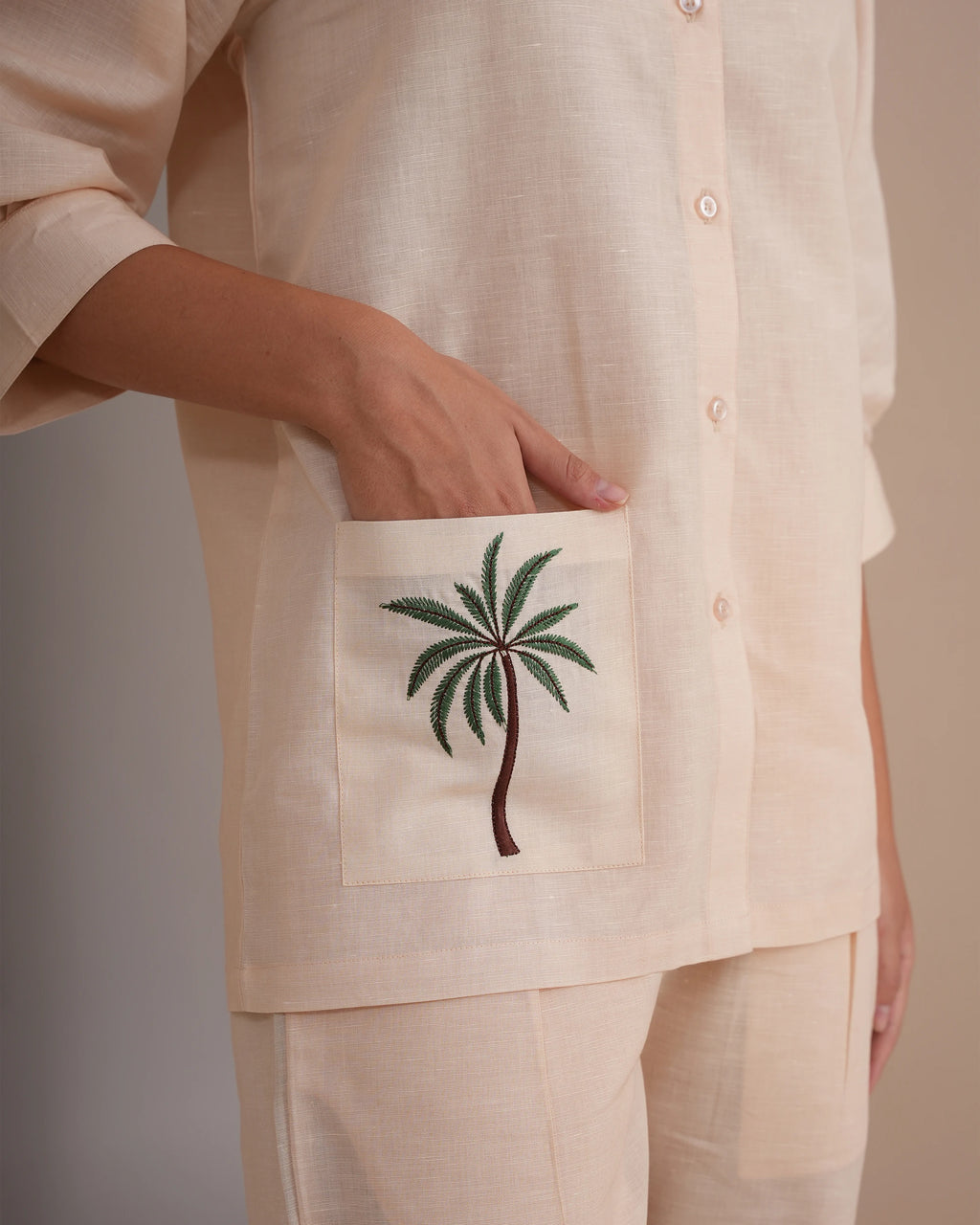 A close-up of a person wearing a light beige linen button-up shirt with a front pocket featuring a green and brown palm tree embroidery, paired with matching beige pants.