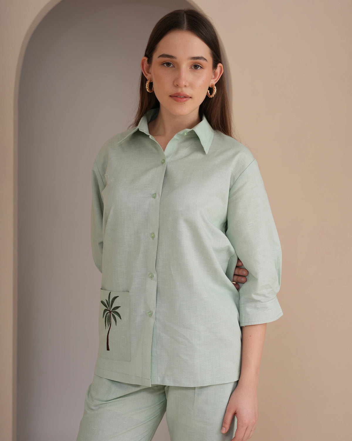 A woman modeling a light sage green linen button-up shirt with a matching pair of trousers. The shirt features a collar, three-quarter sleeves rolled to the forearm, a button-down front, and a pocket on the left side with a black embroidered palm tree motif.