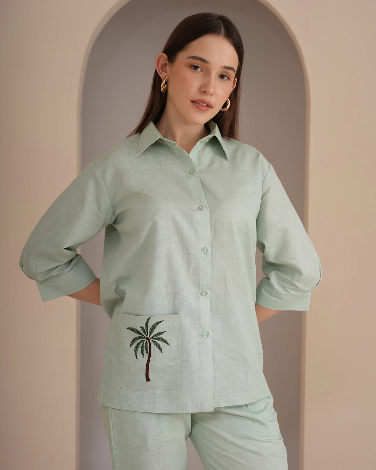 A woman modeling a light sage green linen button-up shirt and matching pants set. The shirt features a chest pocket with an embroidered palm tree design.