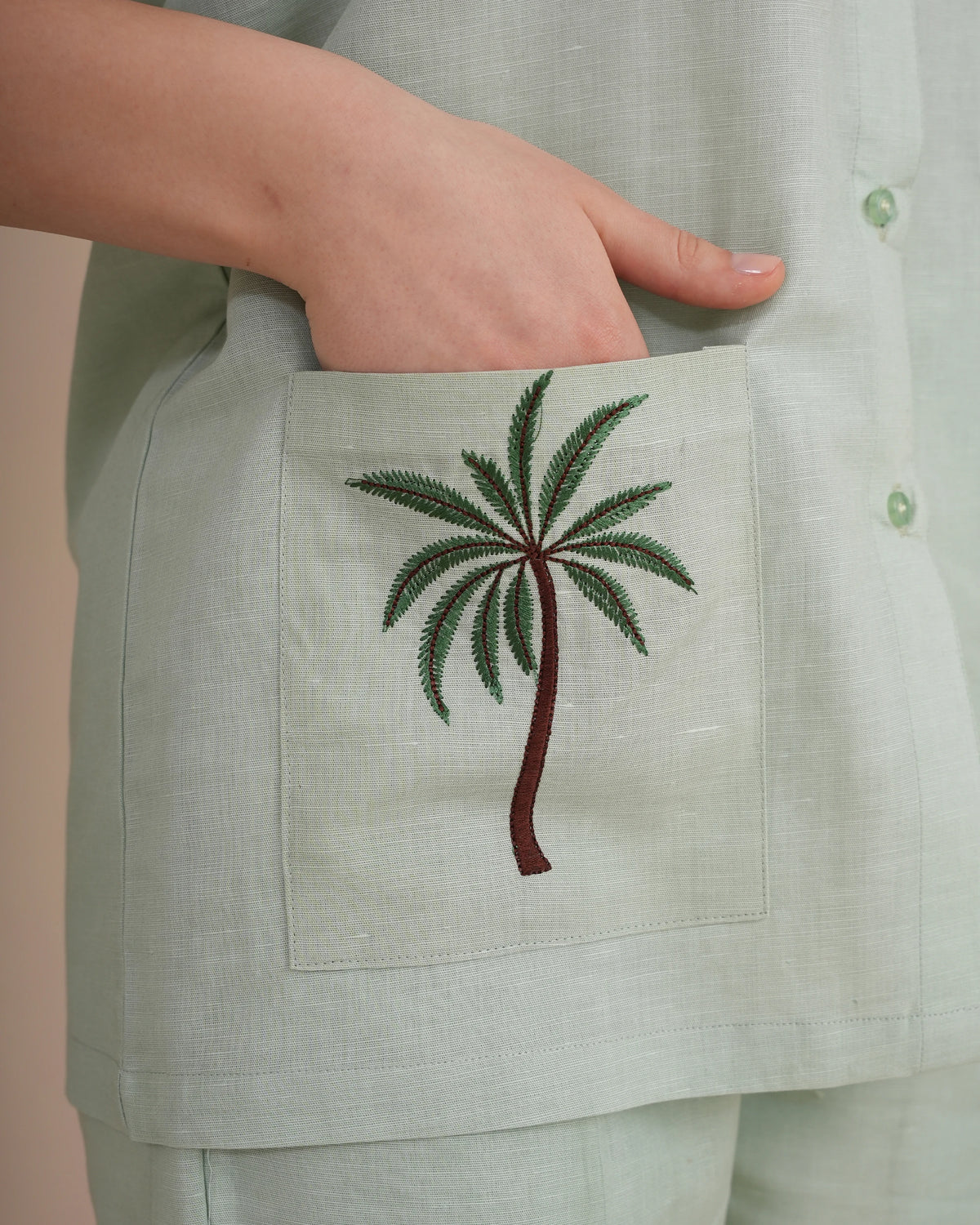 Close-up of a person wearing a light sage green linen button-up shirt with a front pocket featuring an embroidered palm tree motif.
