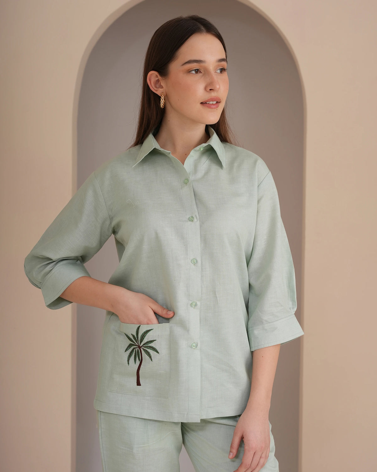 A woman modeling a light sage green linen shirt with a palm tree embroidery on the pocket, paired with matching pants, set against a warm beige arched background.