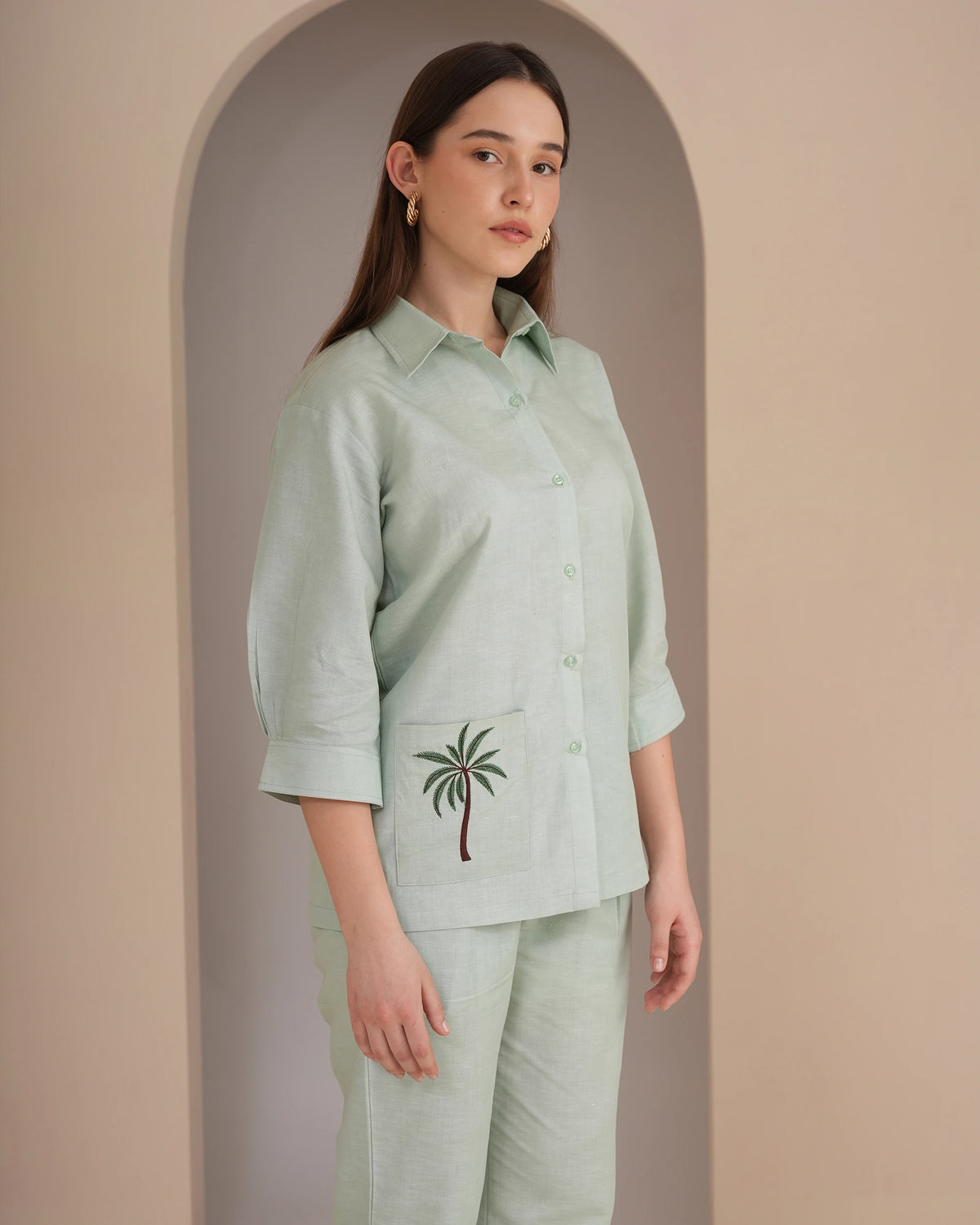 A woman modeling a light sage green linen shirt and matching trousers set. The shirt features a button-down front, three-quarter sleeves rolled to the forearm, and a front pocket decorated with an embroidered green palm tree.