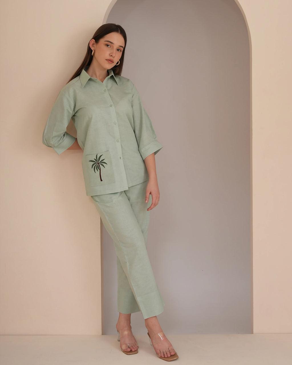 A woman models a light sage green two-piece linen set consisting of a long-sleeve button-up shirt with a palm tree embroidery on the pocket and matching cropped trousers, set against a minimalist beige and gray arched wall.