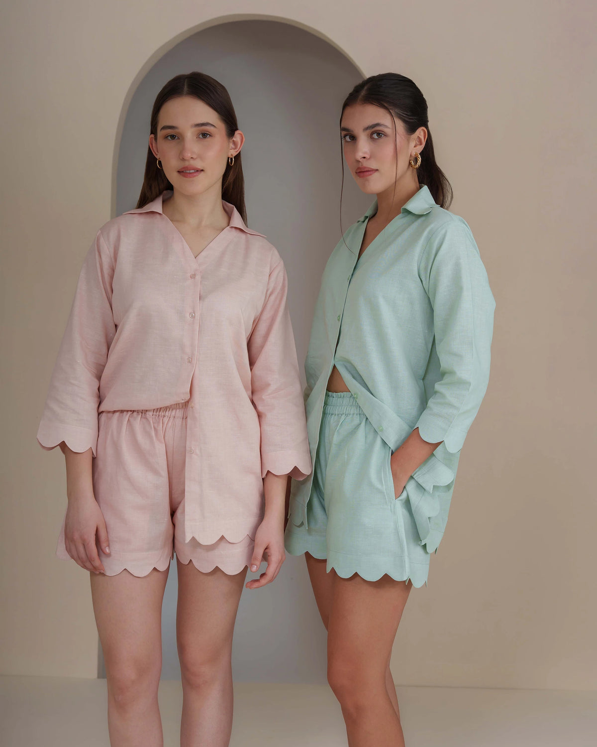 Two women modeling matching two-piece pajama sets with scalloped edges. The set on the left is light pink, and the set on the right is mint green.