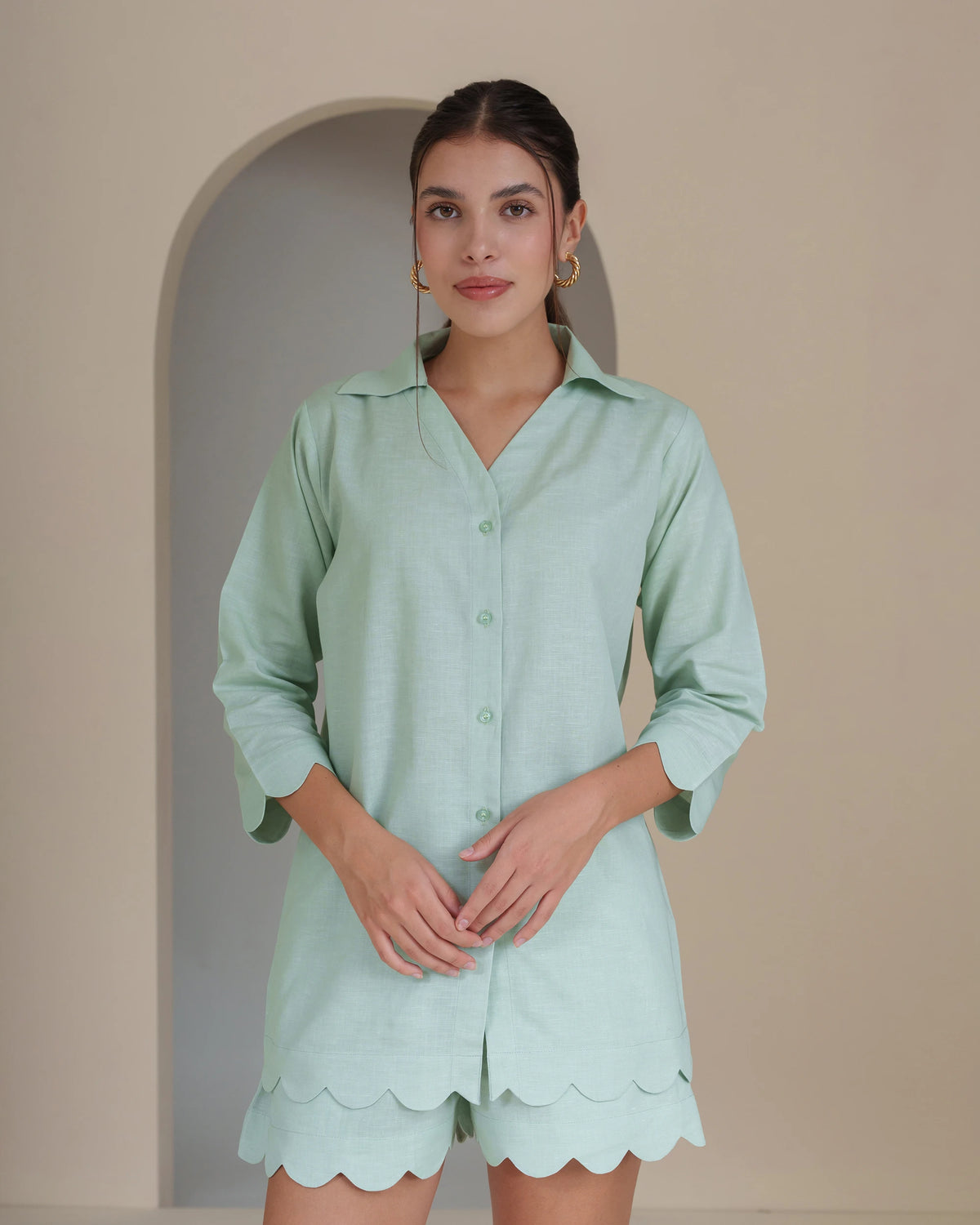 A woman modeling a light mint green short-sleeve button-up shirt with matching scalloped shorts, standing against a beige wall with an arched recess.