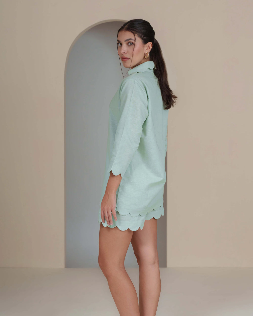 A woman modeling a mint green short-sleeve shirt and matching scalloped mini skirt against a minimalist beige studio backdrop with an arched doorway.