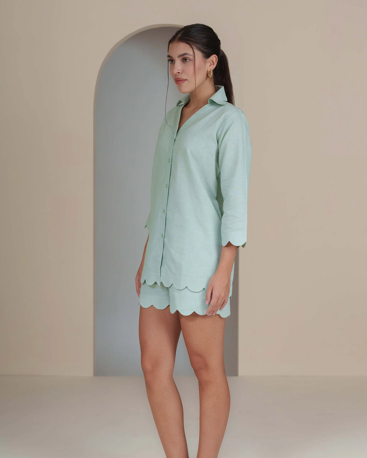 A woman modeling a mint green button-up shirt dress with scalloped edges, standing in front of a minimalist beige wall with an arched alcove.