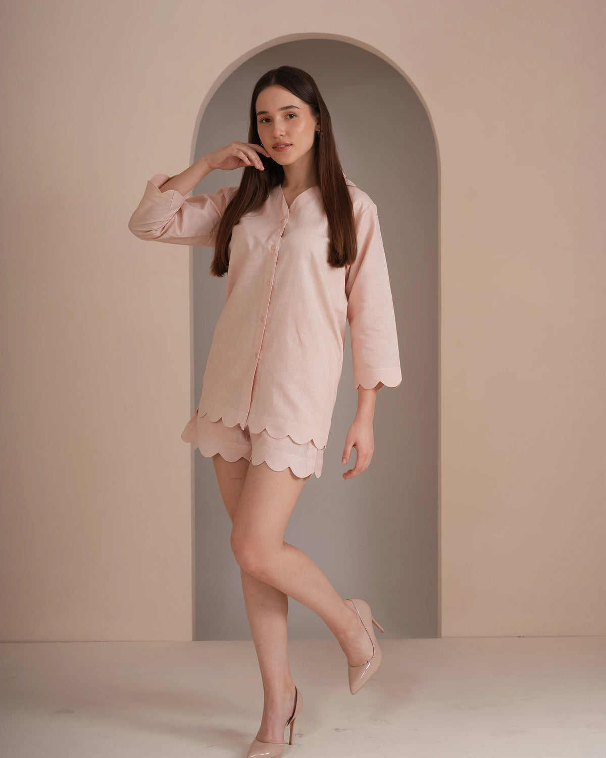 A woman modeling a light pink long-sleeve button-up shirt with a scalloped hem, paired with matching scalloped shorts and nude heels, standing in front of a beige arched wall.