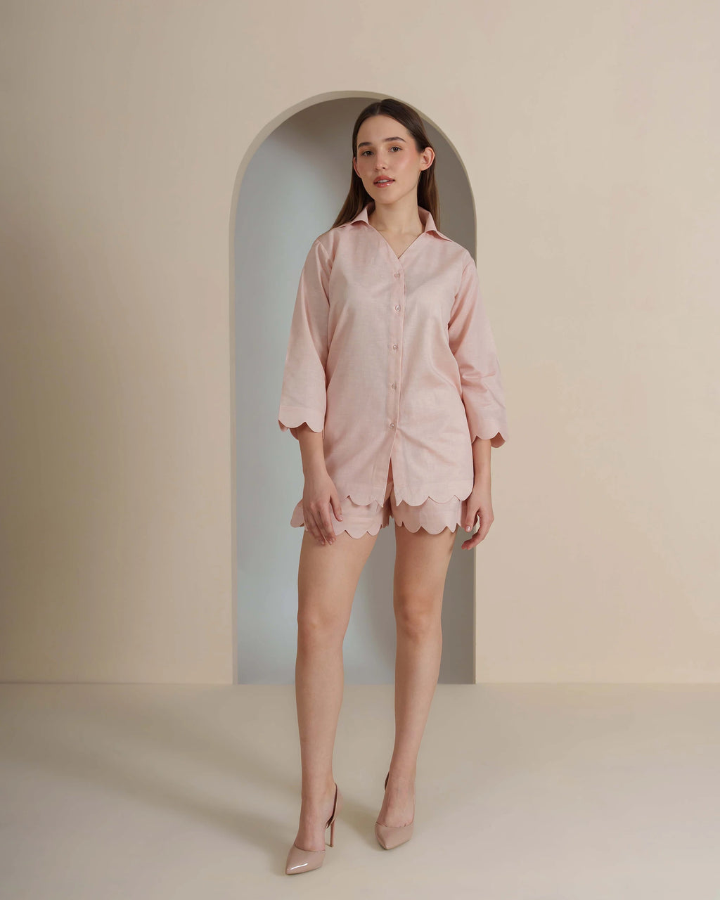 A woman modeling a light pink short-sleeve button-up shirt and matching scalloped shorts set, standing in front of a beige wall with an arched alcove background.