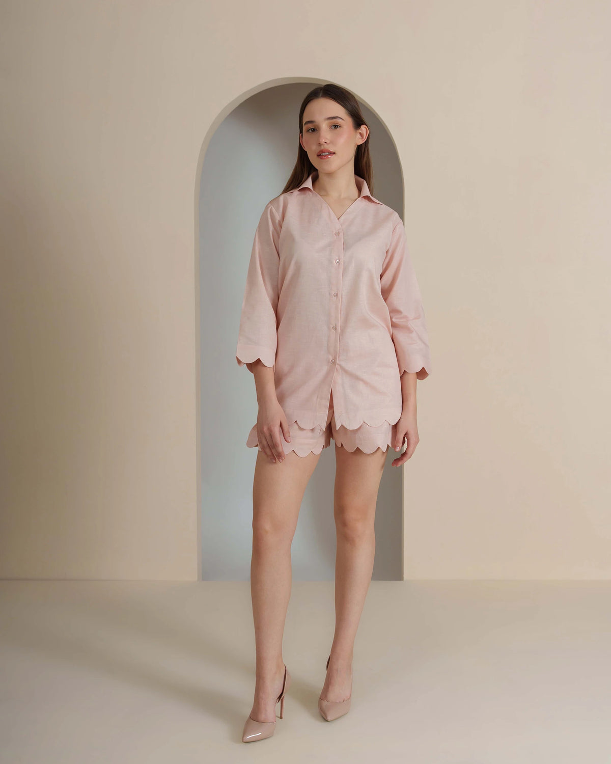 A woman modeling a light pink short-sleeve button-up shirt and matching scalloped shorts set, standing in front of a beige wall with an arched alcove background.