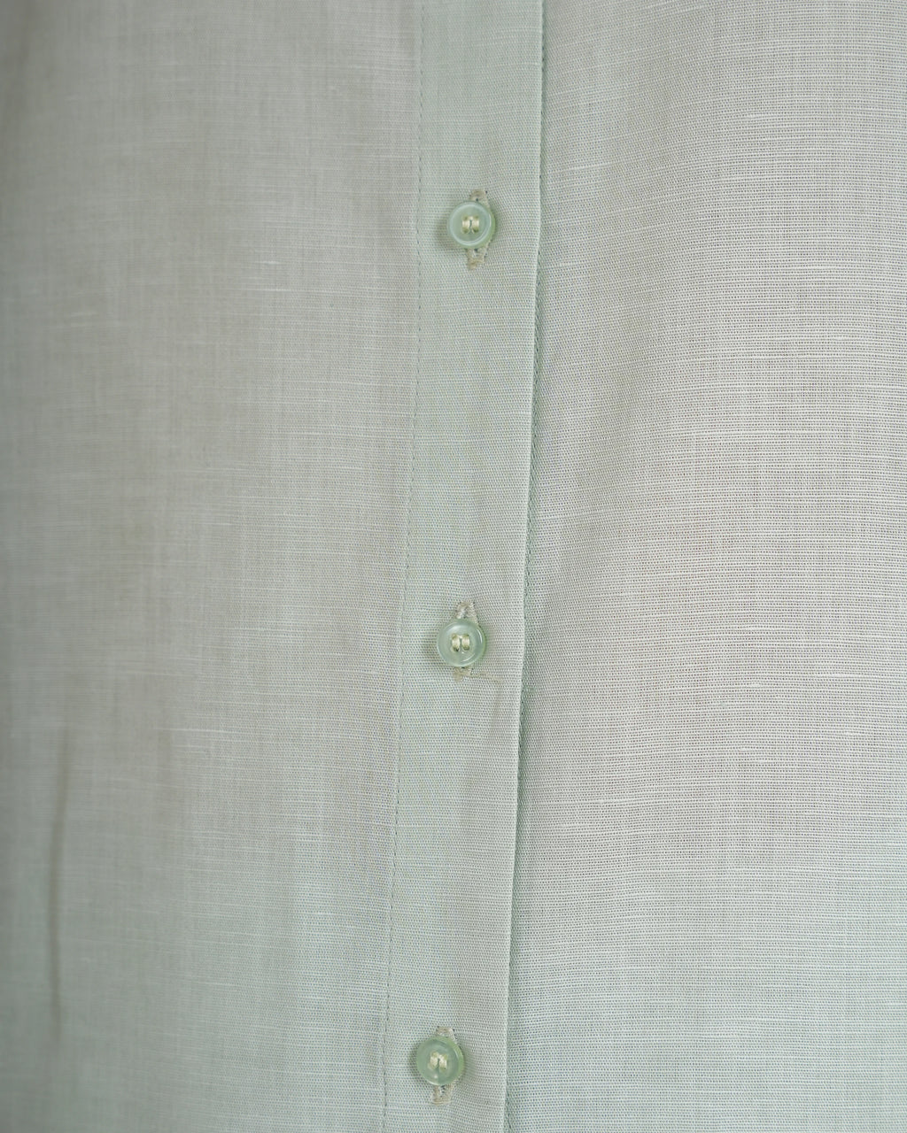 Close-up of a light sage green linen button-up shirt showing the fabric texture and three matching buttons.
