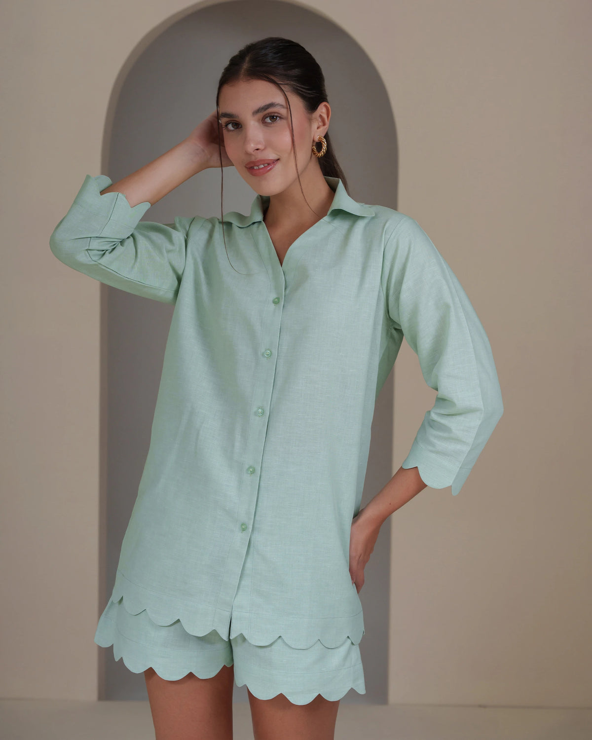 A woman modeling a mint green two-piece lounge set consisting of a long-sleeve button-up shirt and matching scalloped shorts, posed in front of a beige arched wall.
