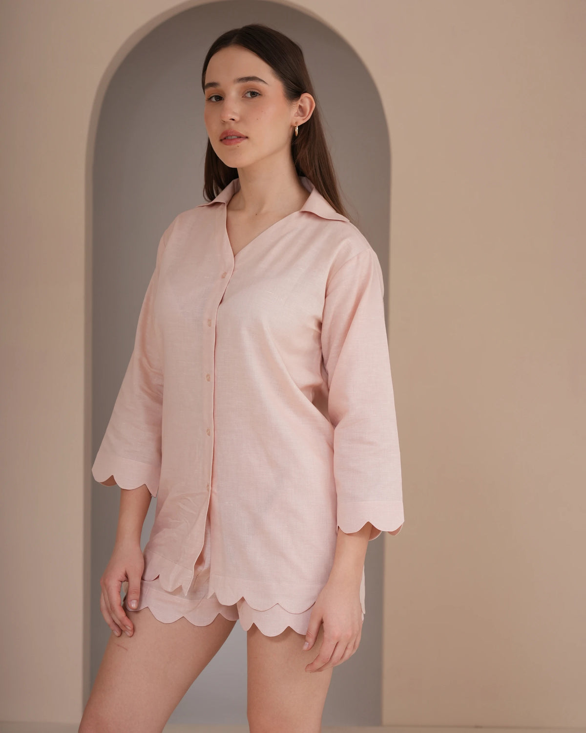 A woman modeling a light pink three-quarter sleeve button-up shirt with scalloped edges, paired with matching pink shorts, set against a warm beige background with an arched alcove.
