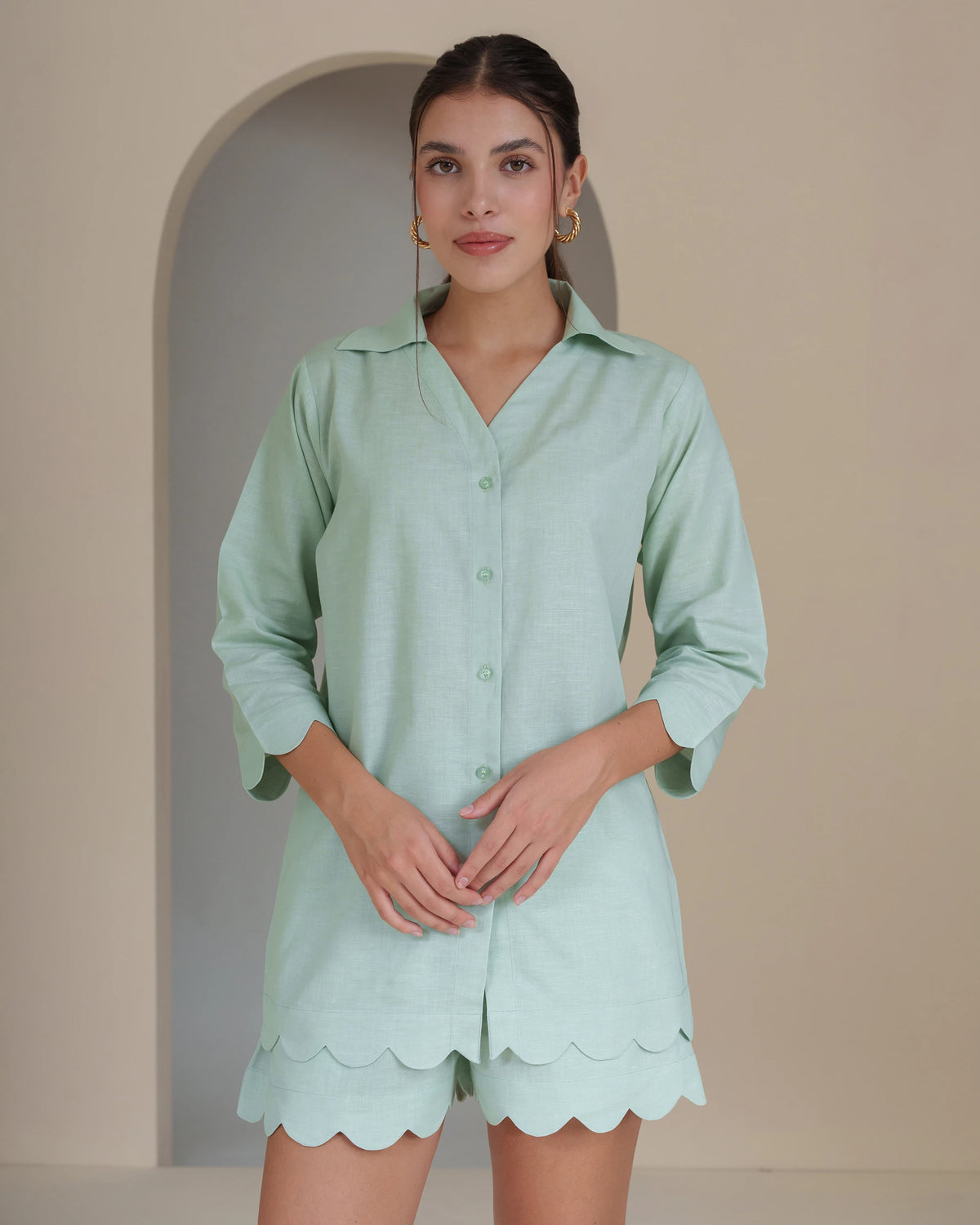 A woman modeling a mint green short-sleeve button-up shirt and matching scalloped shorts set against a beige arched wall background.
