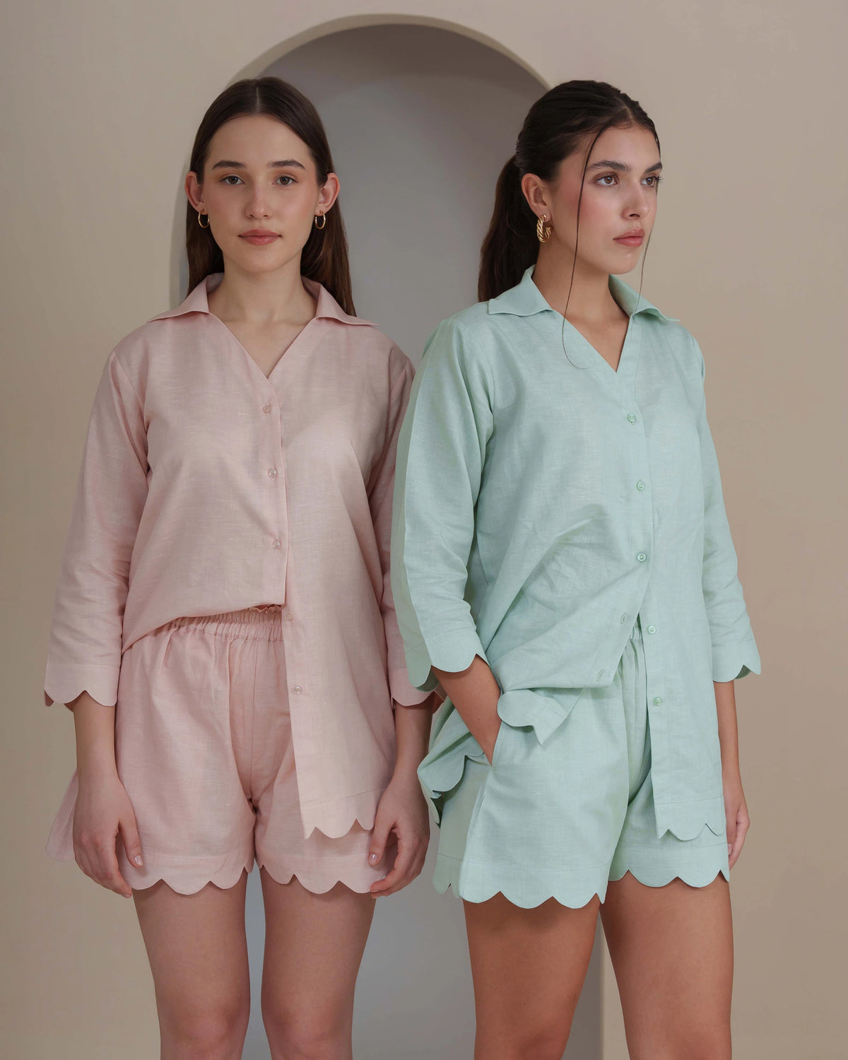 Two women modeling matching pajama sets in a studio setting. The set on the left is light pink, and the set on the right is mint green, both featuring a long-sleeve button-front shirt with scalloped hems and matching shorts.