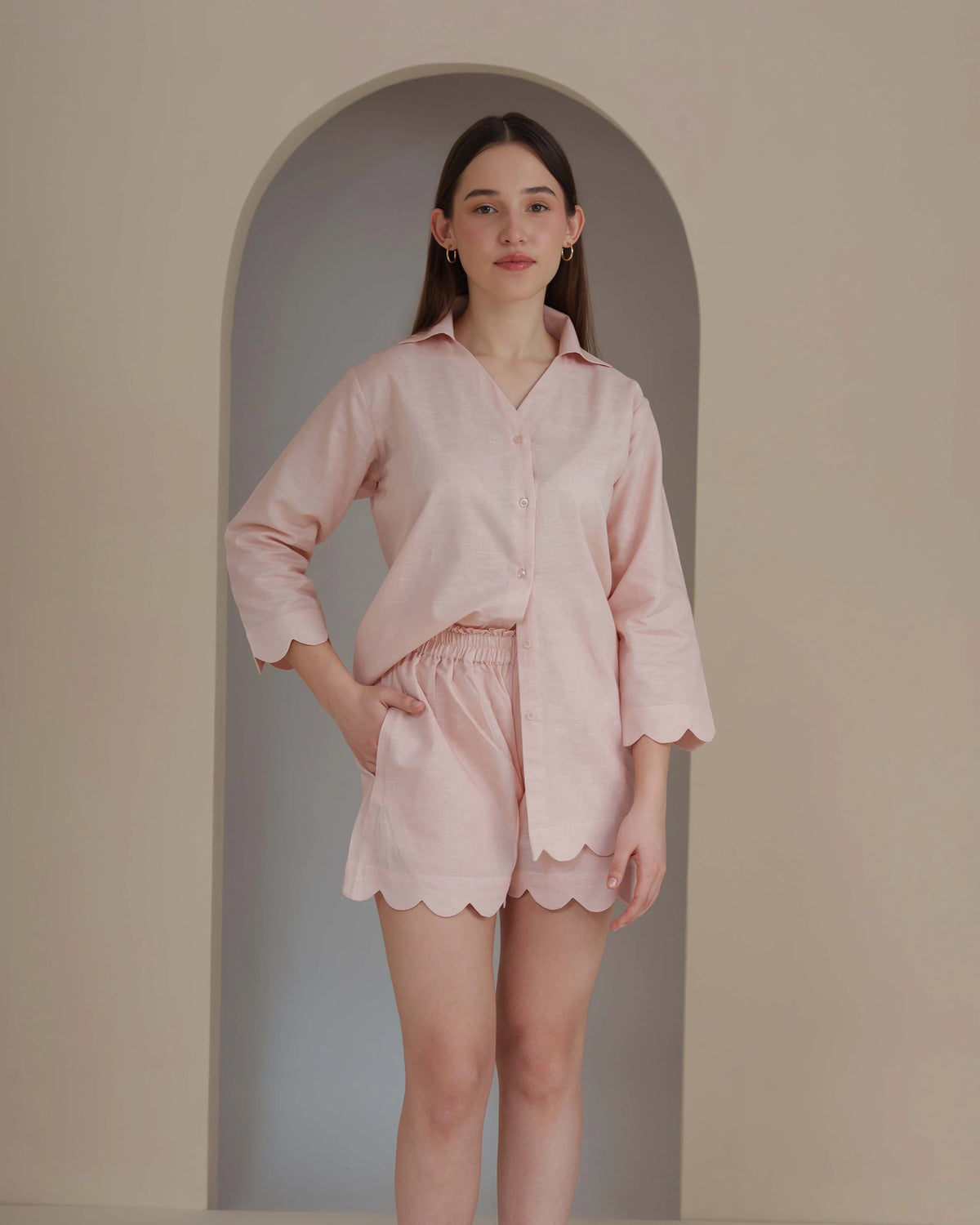 A woman modeling a light pink two-piece pajama set consisting of a long-sleeve button-up shirt and matching shorts with scalloped edges, standing in front of a beige wall with an arched recess.