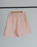 Light pink linen-blend shorts with a scalloped hem and elastic waistband, hanging on a wooden hanger against a light gray background.
